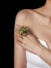 Against a black background, a person in a strapless white top holds the CAMELLIA Taurus Flower Ring—a decorative floral arrangement featuring green and gold elements, possibly with dried preserved flowers—in their left hand.